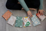Sitting on the floor, a person arranges colorful patterned envelopes and impack.co’s SALE - Mountain Clay Mailers 10" x 13", ensuring each piece blends style with biodegradable transit protection.