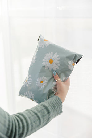 A hand holding a light green package of impack.co's SALE - Daisy White Biodegradable Mailers (10" x 13"), featuring charming white daisy patterns.