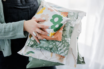 A person holds a stack of vibrant SALE - Daisy Multicolor Biodegradable Mailers 6" x 9" from impack.co, showcasing colorful floral patterns. These mailers provide excellent transit protection, merging style with sustainability in one eco-friendly solution.