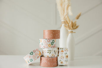 Five rolls of impack.co's SALE - Packing Tape in Daisy Multi print stacked on a white surface, with two white vases and pampas grass in the background, ideal for eco-friendly packaging.