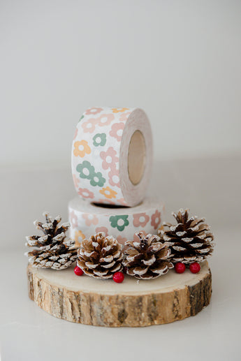 Two floral-patterned rolls of impack.co's "SALE - Packing Tape - Daisy Multi" are stacked on a wooden base, adorned with pine cones and red berries. Perfect for eco-friendly packaging, this water-activated gummed tape combines charm with sustainability.