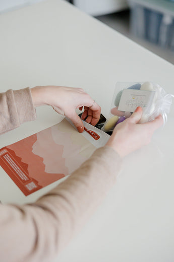 On a white table, someone places colored wool pieces into a plastic bag, preparing to ship sustainably with impack.co's Mountain Clay Mailers 6" x 9", made from biodegradable materials.