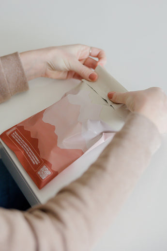 Someone gently opens a SALE - Mountain Clay Mailers 6" x 9" CA from impack.co on a pristine surface, appreciating the elegant design and sustainability of the biodegradable pink and white gradient packaging.