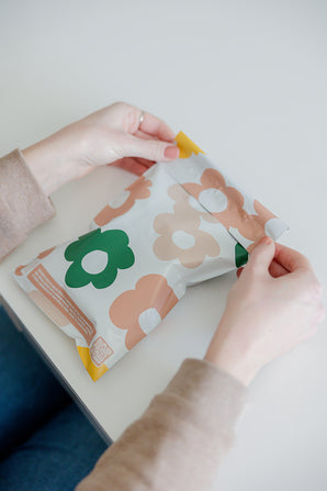 Hands opening an impack.co package with cosmos print on a white surface, protected by SALE - Daisy Multicolor Biodegradable Mailers (6" x 9").