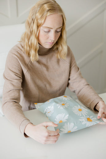 Someone with blonde hair in a beige sweater admires a blue floral-patterned package on a white table, impressed by its design and protection; it's the SALE - Daisy White Biodegradable Mailers 10" x 13" from impack.co.