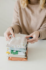 A person in a beige sweater is arranging a stack of colorful Cosmos Biodegradable Mailers by impack.co on a white surface.
