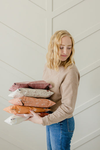 A person in casual attire holds a stack of impack.co's Wildflower Biodegradable Mailers, measuring 10" x 13", against a patterned wall, exuding rustic charm.