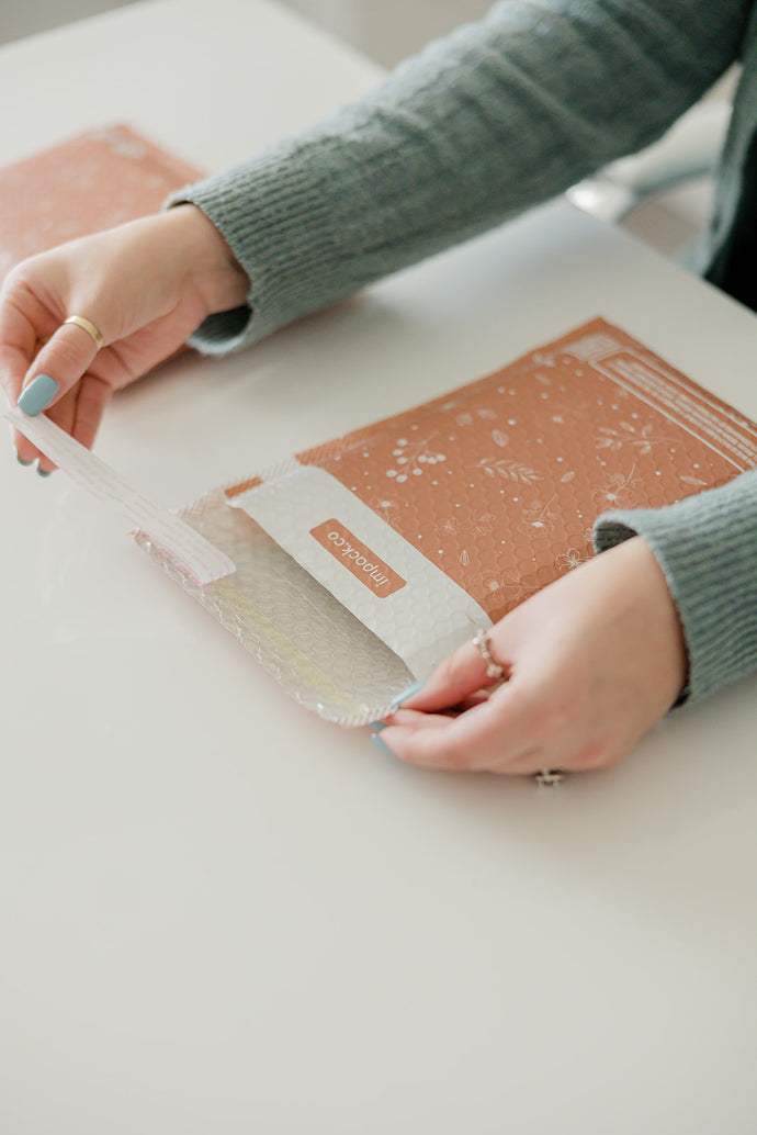 A woman is holding a Rosy Brown Biodegradable Bubble Mailer 6" x 9" from impack.co on a table.