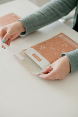 A woman is holding a Rosy Brown Biodegradable Bubble Mailer 6" x 9" from impack.co on a table.