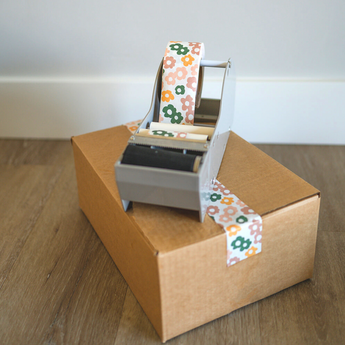 A tape dispenser with impack.co's SALE - Packing Tape in Daisy Multi print sits on a cardboard box, surrounded by its eco-friendly packaging on the wooden floor.